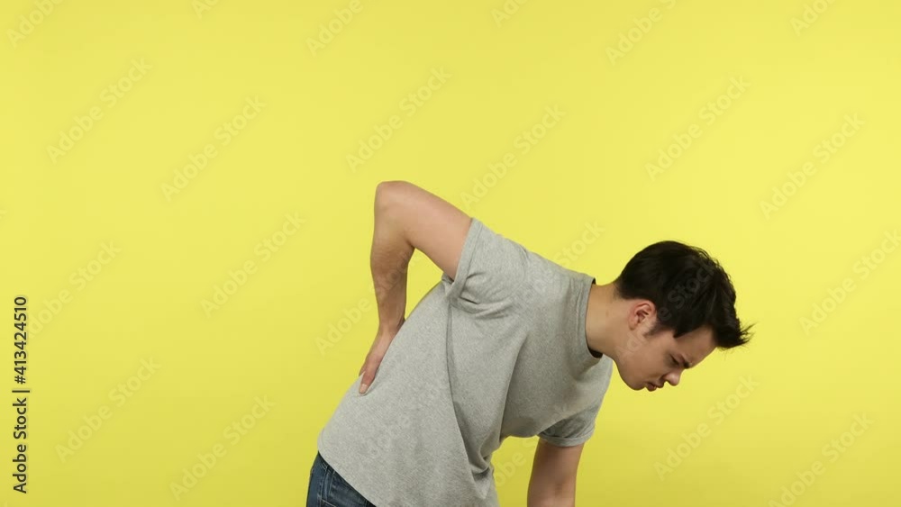 Unhappy overworked young man suffering backache, trying to straighten, touching sore lower spine, unbearable spasm, kidney stones, pinched nerve. Indoor studio shot isolated on yellow background