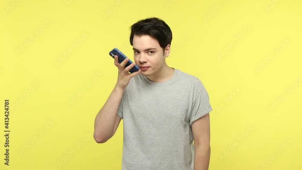 Young teenager in casual style gray t-shirt giving voice command to assistant in his smartphone, searching information using voice instead of typing. Indoor studio shot isolated on yellow background Young teenager in casual style gray t-shirt giving voice command to assistant in his smartphone, searching information using voice instead of typing. Indoor studio shot isolated on yellow background