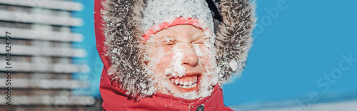 Funny smiling girl in red jacket squinting eyes from bright sun. Happy kid with snow on face at cold winter sunny day. Winter seasonal outdoors fun activity for children. Web banner header.