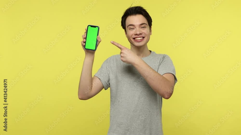Extremely happy positive young man in gray t-shirt pointing finger at green screen smartphone in his hand, boasting with new cool application. Indoor studio shot isolated on yellow background