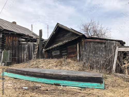 An old rickety wooden hut with boarded-up windows. There is dry grass and trees all around. The house is falling apart from old age, the boards are darkened. An overturned boat is lying in a clearing.
