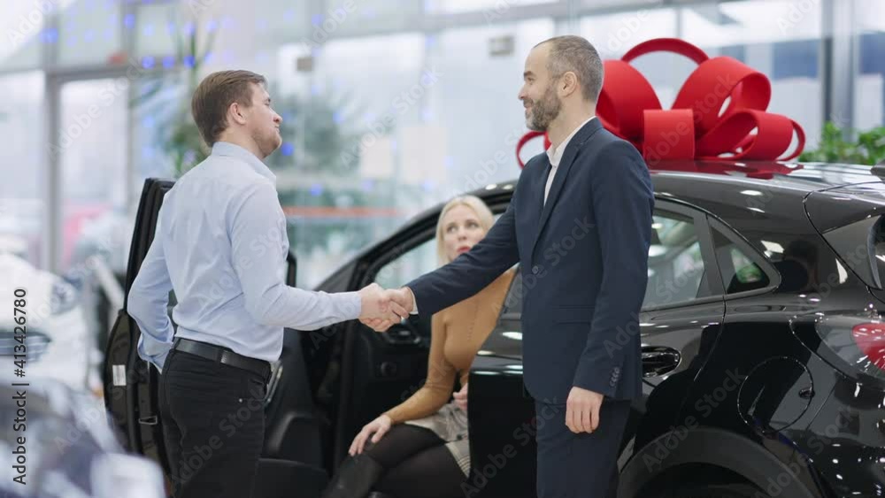 Men shaking hands in car dealership with smiling blurred woman sitting on driver's seat at background. Handshake of confident Caucasian salesman and buyer in showroom. Sale and purchase of vehicle. Men shaking hands in car dealership with smiling blurred woman sitting on driver's seat at background. Handshake of confident Caucasian salesman and buyer in showroom. Sale and purchase of vehicle.