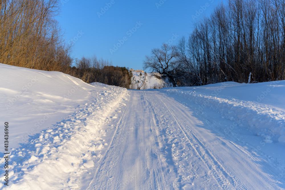 Snow covered road leading to village houses on a high mountain on a frosty winter day. various trees grow along the sides. Pure white snow and pale blue sky Snow covered road leading to village houses on a high mountain on a frosty winter day. various trees grow along the sides. Pure white snow and pale blue sky