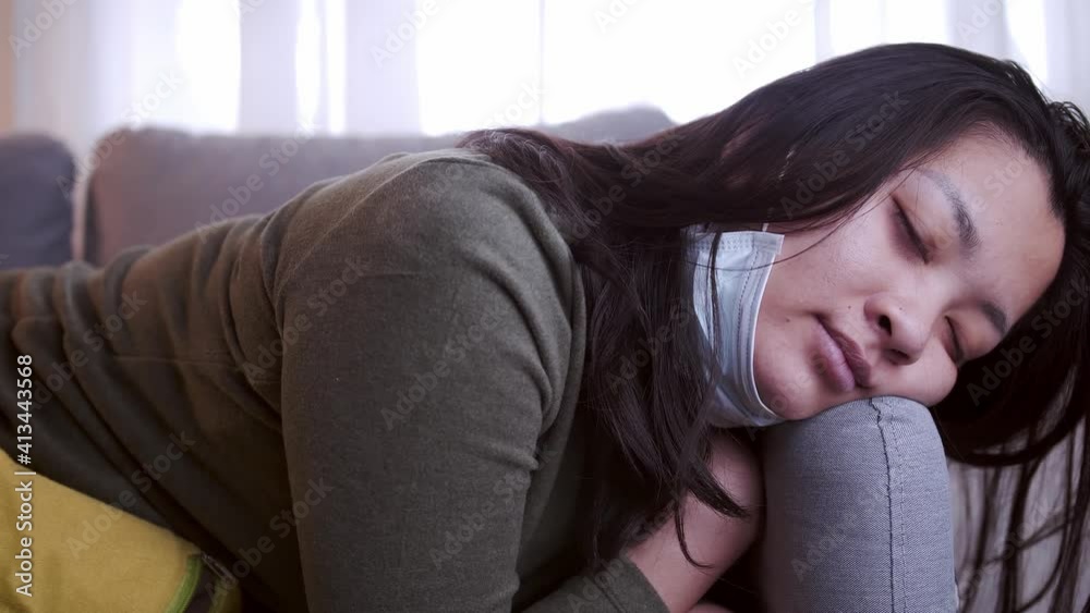 Young Asian woman suffering from coronavirus has black eyes chills all over her body gets cold and uses a mask and blanket to keep warm on the sofa in her separate living room and quarantine Young Asian woman suffering from coronavirus has black eyes chills all over her body gets cold and uses a mask and blanket to keep warm on the sofa in her separate living room and quarantine