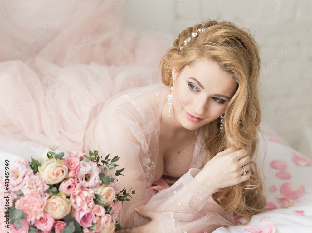 Portrait of a beautiful young girl with long hair in transparent peignoir lies on bed next to bouquet of flowers. Morning of bride
