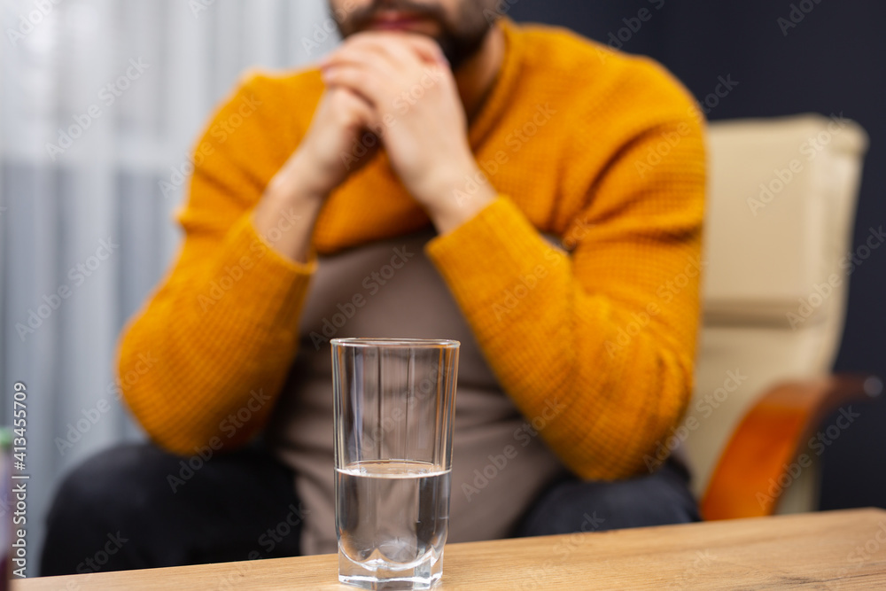 Glass of water on the background of a pensive caucasian man holding his hands under his beard. Indoor shot. Selective focus