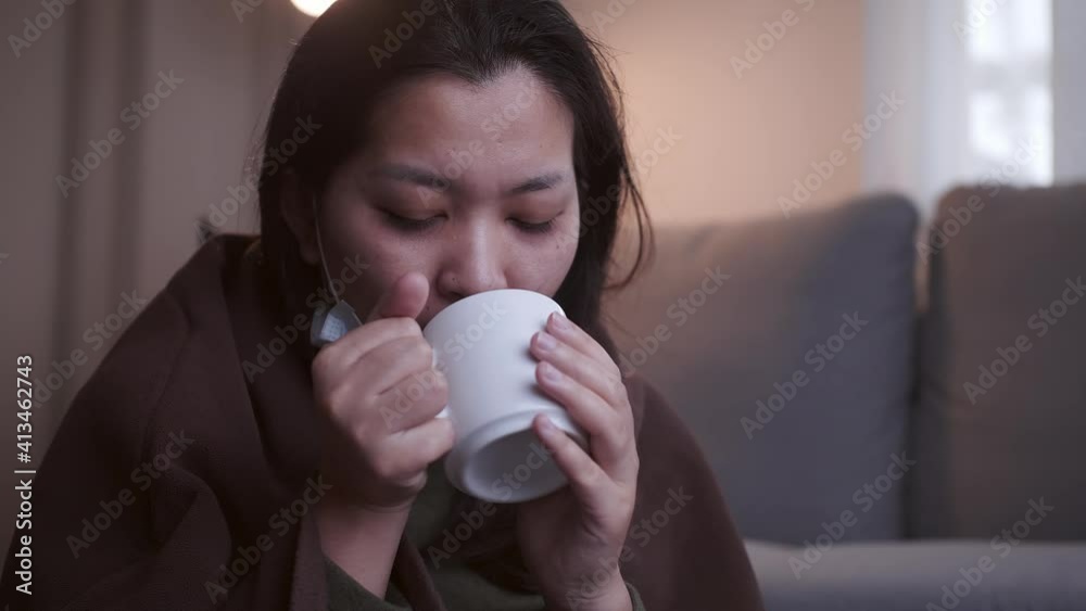 Sick Asian woman drinking lukewarm water in times of suffering has chilling black eyelids all over her body due to a cold and uses masks and blankets to warm and quarantine herself Sick Asian woman drinking lukewarm water in times of suffering has chilling black eyelids all over her body due to a cold and uses masks and blankets to warm and quarantine herself