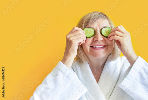 Cheerful senior woman with cucumber slices covering her eyes in front of her eyes on a yellow background. Spa beauty treatments at home, body care concept, organic cosmetics. Natural spa treatment.
