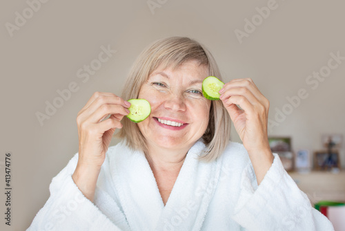 Cheerful senior woman with cucumber slices covering her eyes in front of her eyes in her bedroom. Spa beauty treatments at home, body care concept, organic cosmetics. Natural spa treatment.