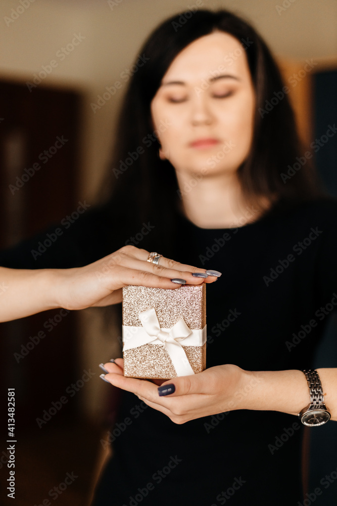 A beautiful brunette girl with long black hair in a black dress holds a golden gift box with a white ribbon bow