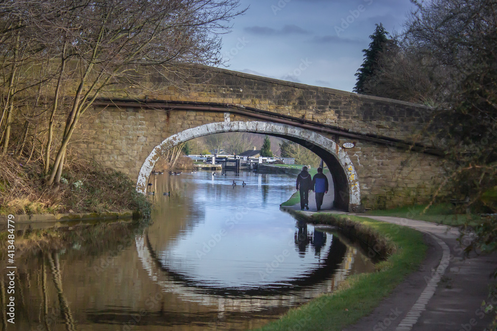 Obraz premium On a bright winter day a couple walk under the bridge on the Leeds and Liverpool Canal at Dowley Gap near Bingley in West Yorkshire