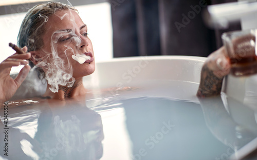 A young sexy tattooed girl enjoying moments of hedonism while having a bath with a cigar and a drink in the bathroom. Relaxation, bath, bathroom, home