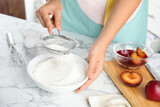 Woman sieving flour at white marble table, closeup. Cooking of delicious plum cake