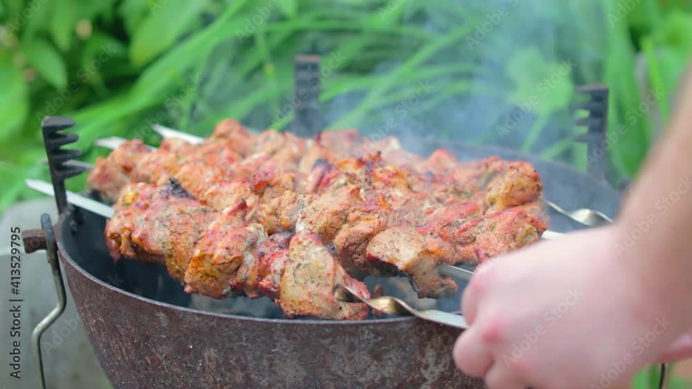 Hot Grilled Kebab Turning on the Mangal in Summer Garden. Delicious Barbecue Meat Grilling on Skewers Against the Green Nature Background. Summer Holiday Picnic Concept - Slow Motion. Middle CloseUp Hot Grilled Kebab Turning on the Mangal in Summer Garden. Delicious Barbecue Meat Grilling on Skewers Against the Green Nature Background. Summer Holiday Picnic Concept - Slow Motion. Middle CloseUp