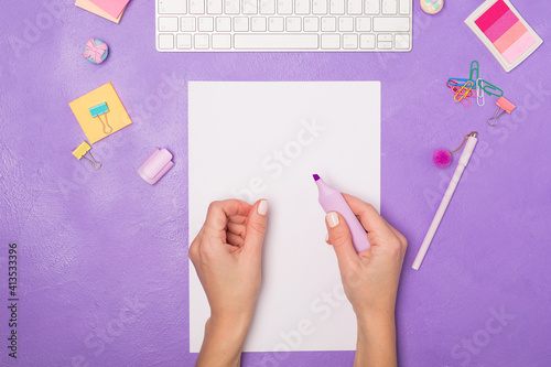 Bright vivid Flat lay workspace. Woman's hand ready for writing on blank list. Items on bright violet purple background, top view. Girly work desktop. School concept. 