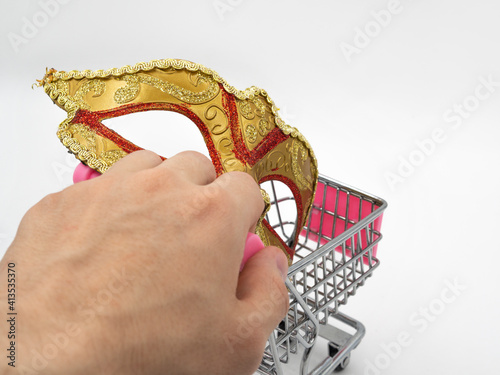 A man holds a toy mini shopping cart (trolley) with a traditional Venetian golden carnival mask on a white background. The concept of preparation for the annual Brazilian festival.