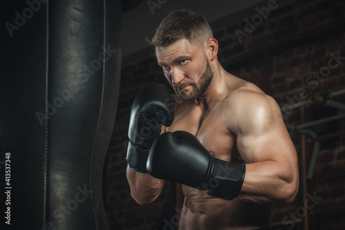 fighter with aggression beats punching bag in black boxing gloves in the gym. boxer training hard, sweat going and skin glittering, copy space © Alexander