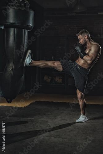 fighter in black boxing gloves beats by leg punching bag in the gym. boxer training hard and with aggression, sweat going and skin glittering, copy space
