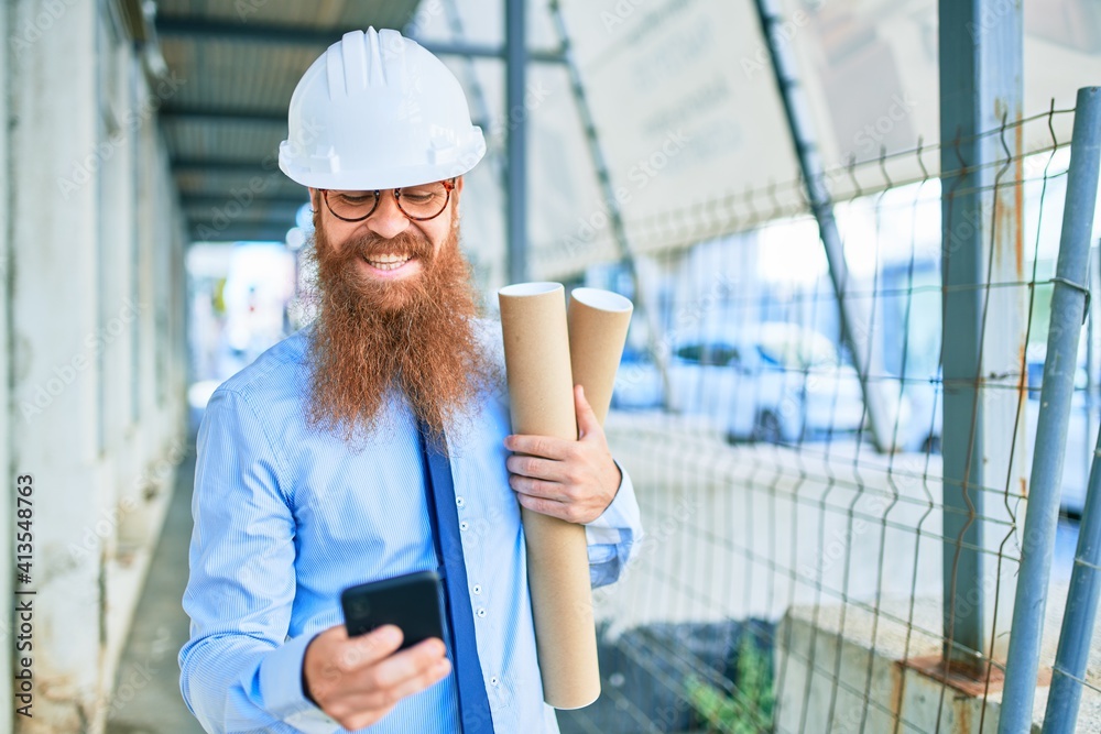 Young redhead architect man with long beard wearing hardhat smiling happy. Holding blueprints using smartphone at street of city. Young redhead architect man with long beard wearing hardhat smiling happy. Holding blueprints using smartphone at street of city.