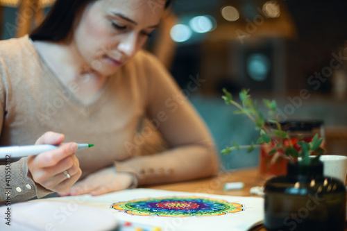 Young woman colouring mandala with markers and white rosary on table with cup of coffee at home photo