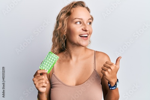 Beautiful caucasian woman holding birth control pills pointing thumb up to the side smiling happy with open mouth