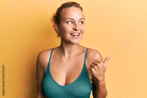 Beautiful caucasian woman wearing sleeveless shirt over yellow background smiling with happy face looking and pointing to the side with thumb up.