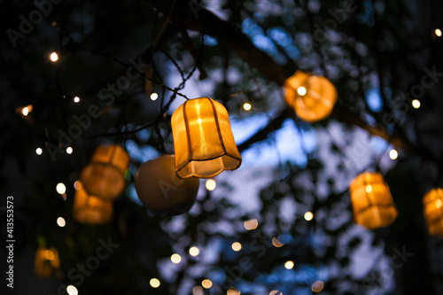 Colorful lampions and lanterns up a tree at night in the garden. A wedding, event or festival banquet decoration at night. Garlands of lamps on a tree branches. Row of paper lampions.