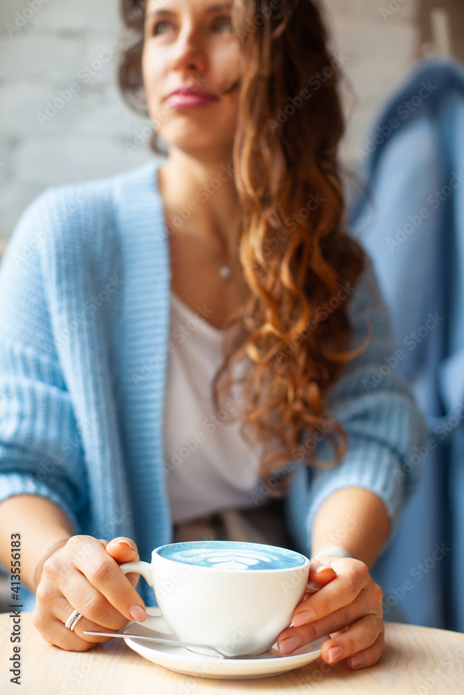 Unfocused happy woman with long wavy hair in warm sweater holding cup of hot blue latte Unfocused happy woman with long wavy hair in warm sweater holding cup of hot blue latte