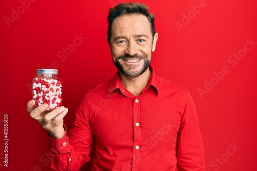 Middle age man holding pills looking positive and happy standing and smiling with a confident smile showing teeth