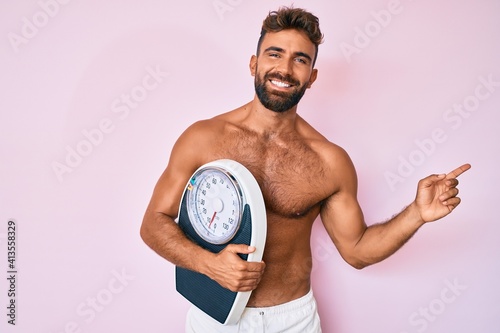Young hispanic man standing shirtless holding weighing machine smiling happy pointing with hand and finger to the side