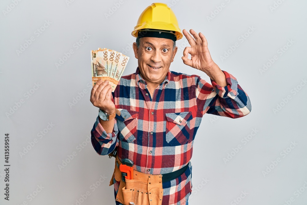 Senior hispanic man wearing handyman uniform holding norwegian krona doing ok sign with fingers, smiling friendly gesturing excellent symbol