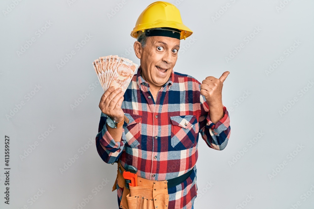 Senior hispanic man wearing handyman uniform holding turkish liras pointing thumb up to the side smiling happy with open mouth