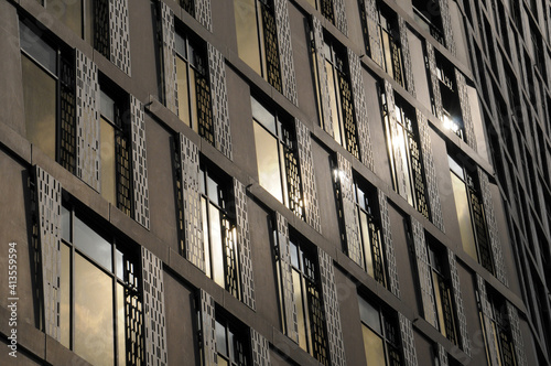 Vue sur une façade métallique brillante bronze et cuivre, vitrages rectangulaires en verre, structures en croix et reflets dorés et blancs du soleil.