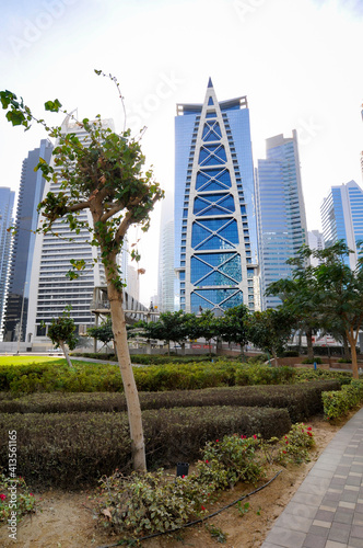 Vue sur un quartier résidentiel et un parc urbain au centre de Dubaï. Verdure, banc, personnes au premier plan et hauts bâtiments à l'architecture moderne en arrière-plan.
