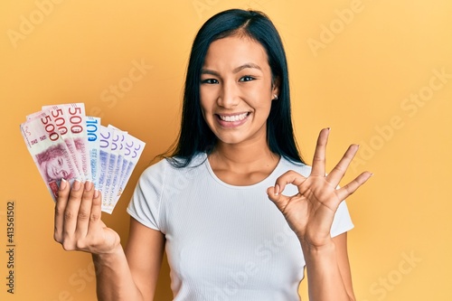 Beautiful hispanic woman holding swedish krona banknotes doing ok sign with fingers, smiling friendly gesturing excellent symbol photo