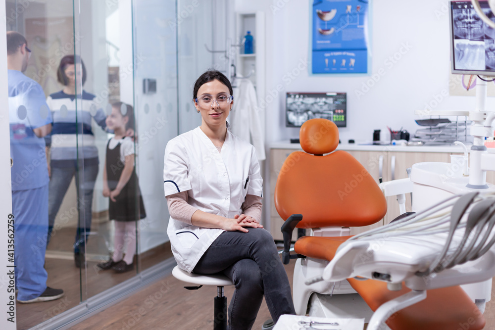 Obraz premium Ortodontist in dentistiry office sitting beside tools tray with assistant discussing with patietns in the background. Stomatolog in professioanl teeth clinic smiling wearing uniform looking at camera.