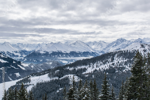 Behind the slope covered with coniferous forest, you can see the panorama of the Kitzbuhel Valley topped by a mountain range with sharp-pointed snow-capped peaks. © Сергей Дворецкий