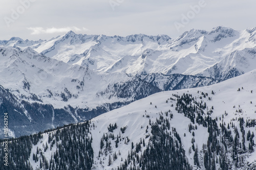 Behind the slope covered with coniferous forest  you can see the panorama of the Kitzbuhel Valley topped by a mountain range with sharp-pointed snow-capped peaks.