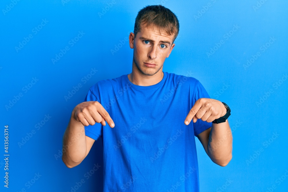Young caucasian man wearing casual blue t shirt pointing down looking sad and upset, indicating direction with fingers, unhappy and depressed. Young caucasian man wearing casual blue t shirt pointing down looking sad and upset, indicating direction with fingers, unhappy and depressed.