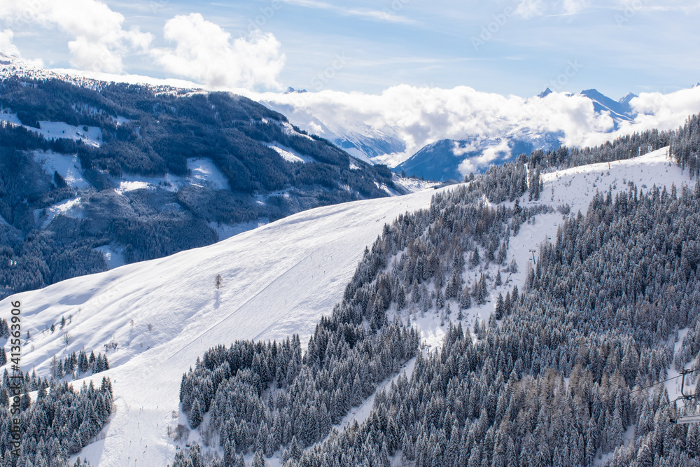 The Austrian Alps in winter near Kitzbuhel. Behind the snow-covered fir trees, there is a perfectly prepared ski track, above which the magnificent mountain peaks rise.