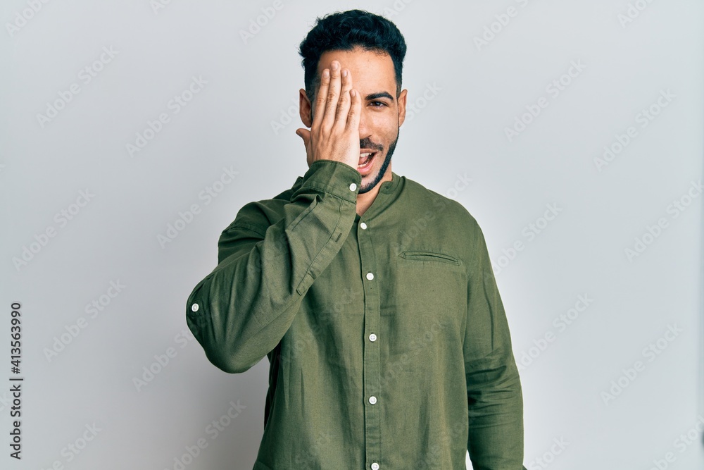 Young hispanic man wearing casual clothes covering one eye with hand, confident smile on face and surprise emotion. Young hispanic man wearing casual clothes covering one eye with hand, confident smile on face and surprise emotion.