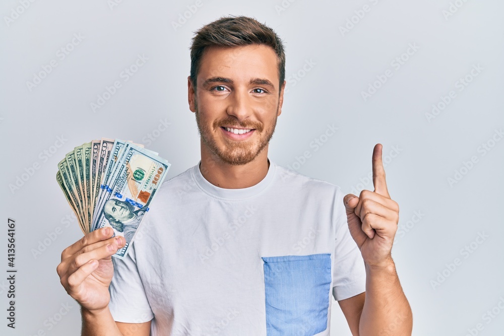 Handsome caucasian man holding dollars smiling with an idea or question pointing finger with happy face, number one Handsome caucasian man holding dollars smiling with an idea or question pointing finger with happy face, number one