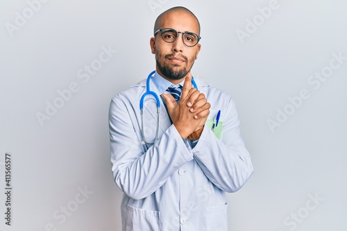 Hispanic adult man wearing doctor uniform and stethoscope holding symbolic gun with hand gesture, playing killing shooting weapons, angry face photo
