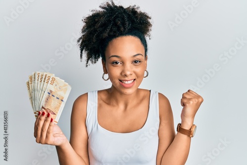 Young african american girl holding 100 danish krone banknotes screaming proud, celebrating victory and success very excited with raised arm photo