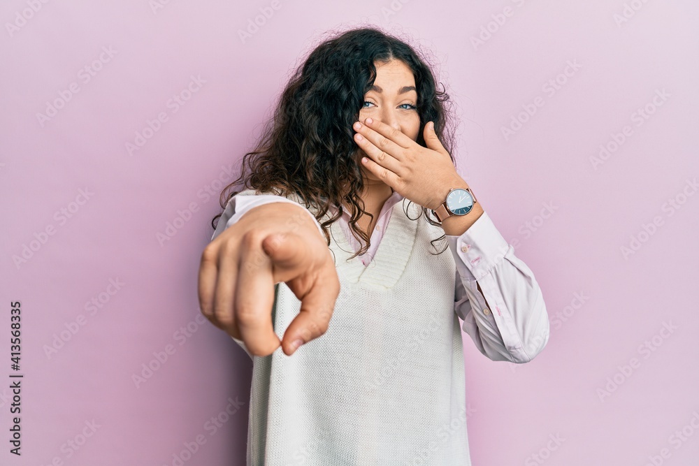 Young brunette woman with curly hair wearing casual clothes laughing at you, pointing finger to the camera with hand over mouth, shame expression