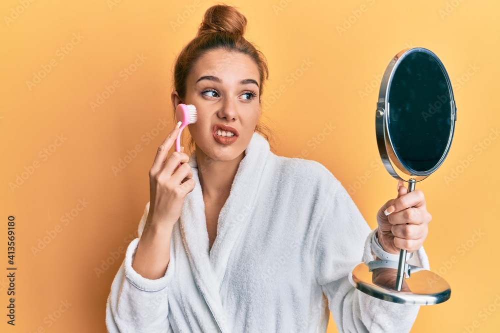 Young blonde woman wearing robe holding makeup remover brush in shock face, looking skeptical and sarcastic, surprised with open mouth Young blonde woman wearing robe holding makeup remover brush in shock face, looking skeptical and sarcastic, surprised with open mouth