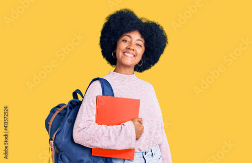 Young african american girl wearing student backpack holding book looking positive and happy standing and smiling with a confident smile showing teeth