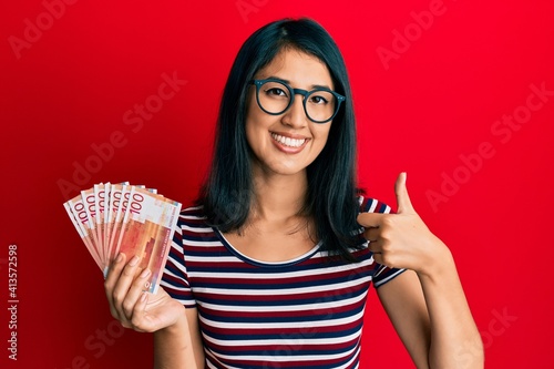 Beautiful asian young woman holding 100 norwegian krone banknotes smiling happy and positive, thumb up doing excellent and approval sign photo