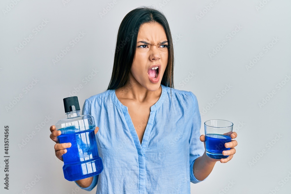 Young hispanic girl holding mouthwash for fresh breath angry and mad screaming frustrated and furious, shouting with anger. rage and aggressive concept. Young hispanic girl holding mouthwash for fresh breath angry and mad screaming frustrated and furious, shouting with anger. rage and aggressive concept.