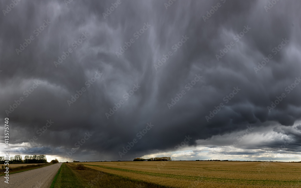 Dramatic blue-gray storm clouds over a panoramic prairie landscape with a gravel road extending off into the distance.
